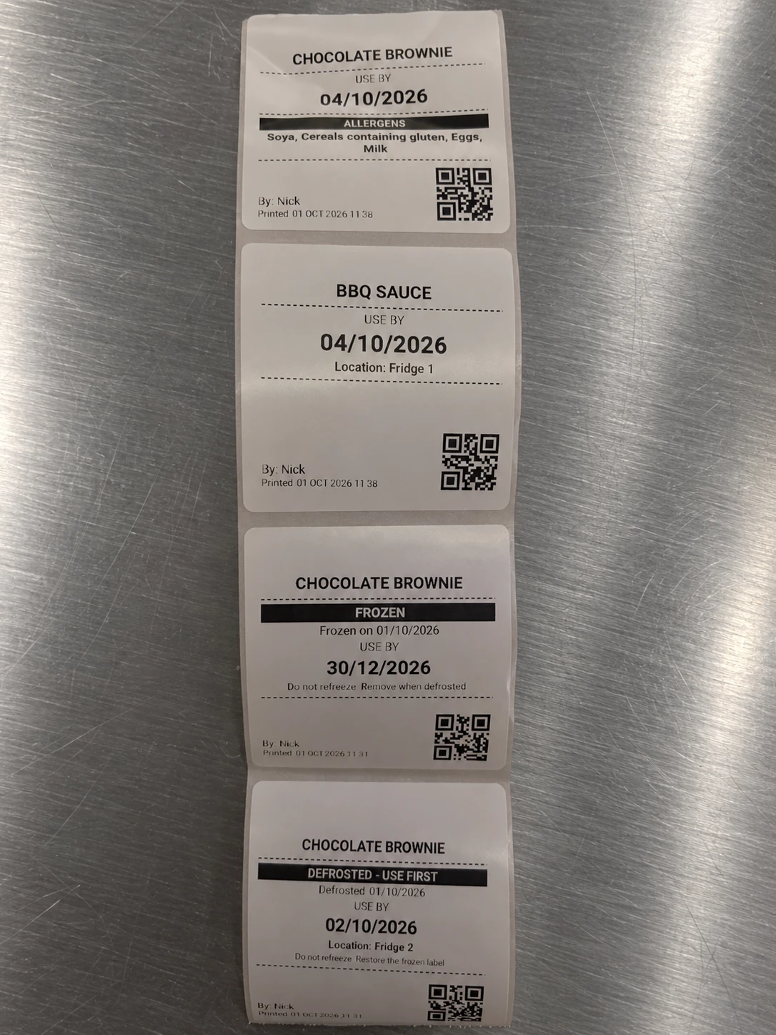 A strip of real CompliChef labels: an allergen label for chocolate brownie, a use-by label for BBQ sauce, and frozen and defrosted labels with use-by dates